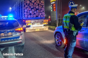 Radiowóz z włączonymi sygnałami świetlnymi, obok policjant kontroluje trzeźwość kierowcy. W tle odjeżdżający samochód ciężarowy z drewnem.