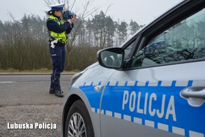 napis policja na radiowozie, w tle policjantka kontroluje prędkość pojazdów